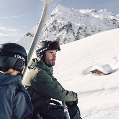 Zwei Skifahrer, eine Frau und ein Mann, sitzen gemeinsam in einem Sessellift und fahren den verschneiten Berg hinauf. Beide tragen Skibekleidung, Helme und Skibrillen. Im Hintergrund erstreckt sich eine tiefverschneite Winterlandschaft mit einem kleinen, fast eingeschneiten Holzhaus und einem imposanten, schneebedeckten Bergmassiv, der Madrisa. Die Stimmung ist ruhig und sonnig, der Himmel klar. | © Gargellner Bergbahnen GmbH & CO KG, Daniel Zangerl